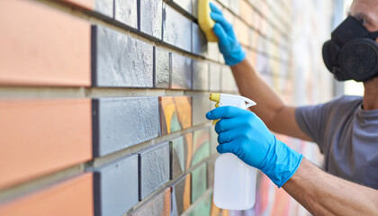 Worker in protective mask, gloves scrubs graffiti from brick wall using cleaning spray, sponge. Professional cleaning service restores building facade. Hygiene, sanitization for outdoor surfaces.