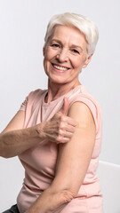 Senior woman wearing face mask showing vaccinated arm with adhesive bandage plaster and gesturing thumbs up. Elderly patient approving vaccination. Covid-19 Vaccination program concept