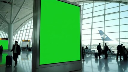 Blank green screen billboard in a modern airport terminal with travelers and an airplane visible through the window. Digital advertising space. - Powered by Adobe