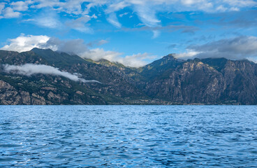 Dramatic mountains surround Lake Garda under a cloudy sky, with sunlight breaking through to highlight the rocky landscape. The calm water contrasts with the powerful peaks of northern Italy.
