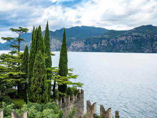 Ancient castle walls rise above Lake Garda, surrounded by tall cypress trees and lush greenery. The view opens to dramatic cliffs and blue water under a cloudy sky in northern Italy.