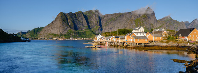 Bright colors of historic wooden cabins line the peaceful waterfront of Sakrisoy in Lofoten, Norway. Majestic mountain peaks rise dramatically under a clear blue sky, creating a serene atmosphere.