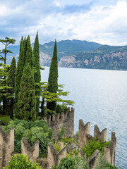 Ancient castle walls rise above Lake Garda, surrounded by tall cypress trees and lush greenery. The view opens to dramatic cliffs and blue water under a cloudy sky in northern Italy.