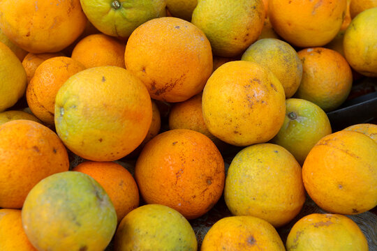 Tight composition of market oranges piled in a shallow crate shows mottled yellow‑orange rinds, natural scuffs, and patches of green. Soft daylight from above reveals pores and pithy scars,