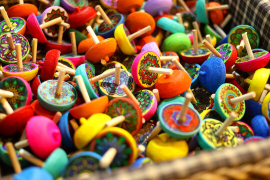 A basket overflows with ornate hand‑decorated spinning toys&mdash;whirligigs rendered in saturated neons and intricate patterns. Lacquered finishes and tiny axles catch glints of light, while curved rims