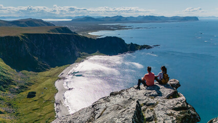 Fototapeta premium Two hikers admire the stunning panorama of the coast from Matind Mountain in Bleik, Andoya. The sun glistens on the ocean waves, highlighting the beautiful landscape and serene atmosphere.