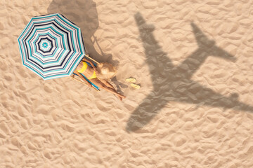 Woman resting under striped umbrella on beach, top view. Flying airplane casting shadow on sand