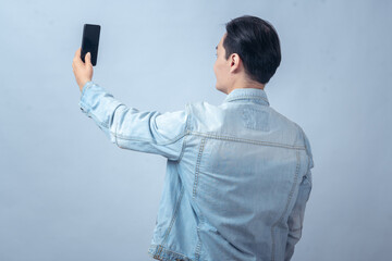 Asian man in denim jacket holding smartphone while facing away, posing as if taking a selfie or...