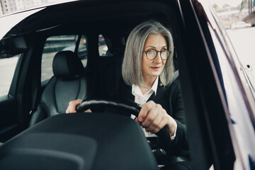 Elegant mature businesswoman driving a car through an urban city street in daylight wearing formal attire