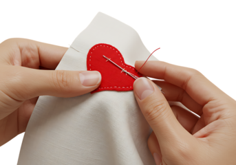 Hands carefully sewing a red heart onto white fabric with needle and thread, creating a loving and handmade craft project in a bright studio.