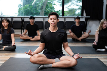 Young fit multiracial people in sportswear sitting in row on floor mat in lotus padmasana position, meditating with closed eyes and folded in mudra gesture fingers on knees at yoga group training.