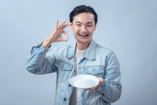 Asian man in denim jacket smiling while holding an empty white plate and giving a thumbs up gesture, isolated on plain studio background - Powered by Adobe