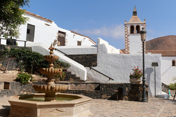 Traditional architecture in Betancuria, Fuerteventura. A stone fountain, whitewashed walls, and the church tower stand under a clear blue sky in this historic Canarian village.