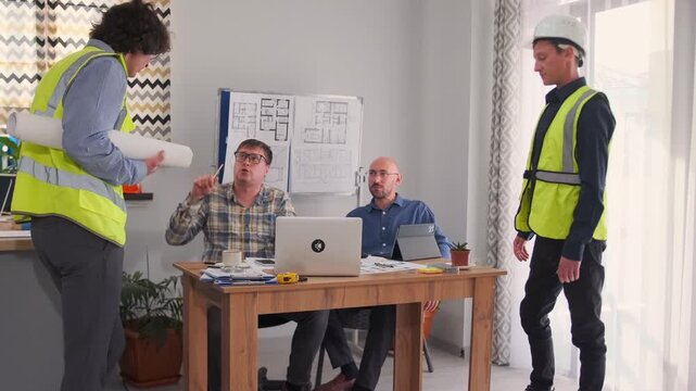 Two construction workers with blueprints argue with architect and project manager seated at office desk. Tense discussion with gestures about building approval and planning.