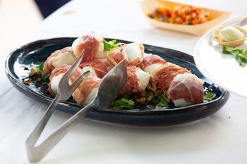 Delicious mozzarella balls wrapped in prosciutto with serving tongs on a plate