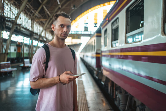 traveler guy boarding train and messaging with phone at busy railroad station, young man with backpack and smartphone starting journey on railway platform