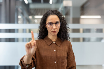 Woman standing in an office, wearing glasses, and showing a stop gesture with her raised index finger, conveying disapproval, rejection, or a serious warning