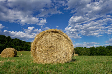 Rolling hay bales sit peacefully in a green field under a bright blue sky filled with clouds.