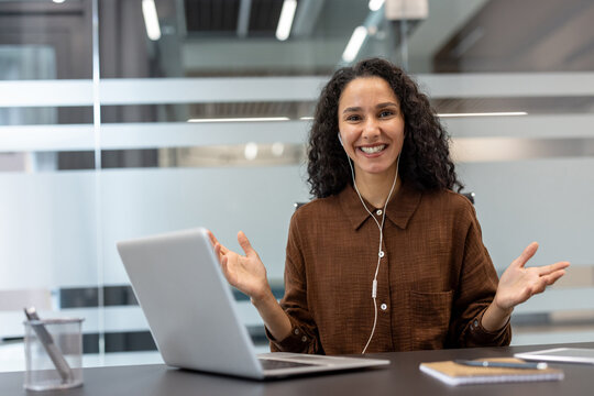 Young businesswoman smiling and gesturing during a virtual meeting, wearing earbuds while presenting and communicating on her laptop in a modern home office workspace - Powered by Adobe