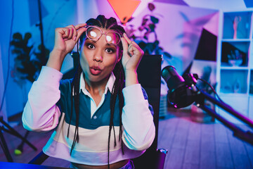Podcaster woman in a neon-lit room adjusts her glasses while hosting a professional podcast or vlog, focusing on media communication.