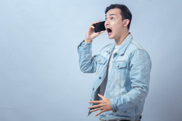 Asian man in denim jacket talking on phone with shocked expression, mouth open wide, showing surprise and disbelief against plain studio background