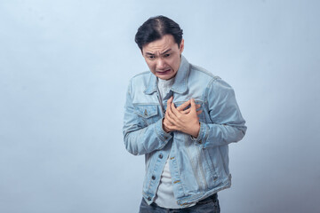 Asian man in denim jacket clutching chest with worried painful expression, standing in studio against plain light background, showing chest pain, heart problem, and discomfort