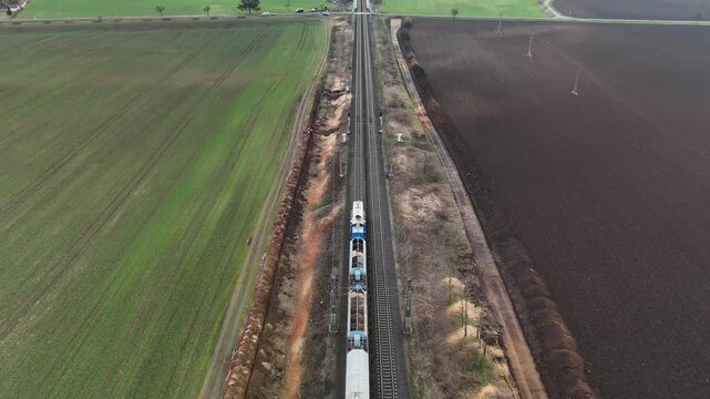 Aerial drone view of a diesel freight train running straight across fields with mountains beyond, highlighting railway logistics, locomotive power, and a calm rural landscape.