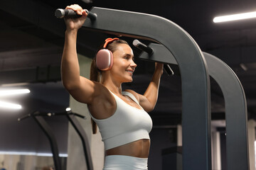 Smiling woman in headphones training on pull up machine in gym