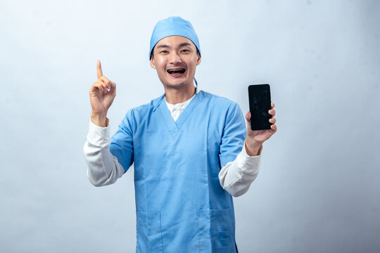 Asian man in blue medical scrubs smiling while holding a smartphone in one hand and pointing upward with the other against a plain studio background