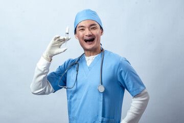 Asian doctor in blue scrubs with stethoscope and gloves holds a syringe confidently, showing...