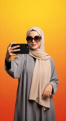 Obraz premium Fashionable woman wearing a hijab and sunglasses taking a selfie with her smartphone, isolated on a vibrant gradient background
