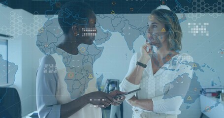 Discussing data businesswomen wearing suits using tablet at office desks, with map overlay monitors