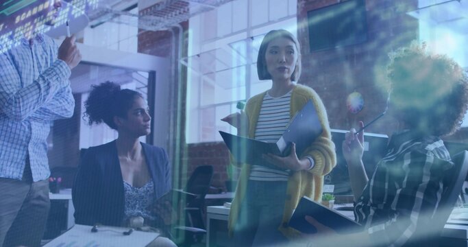 Presenting woman leading team meeting in open-plan office, with open binder and monitors