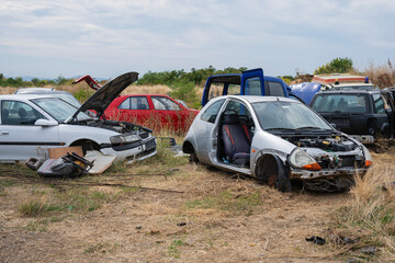Abandoned vehicles in various states of disrepair scattered across a junkyard under a cloudy sky.