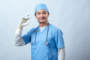 Asian doctor in blue scrubs with stethoscope and gloves holds a syringe confidently, showing determination and readiness against a plain studio background