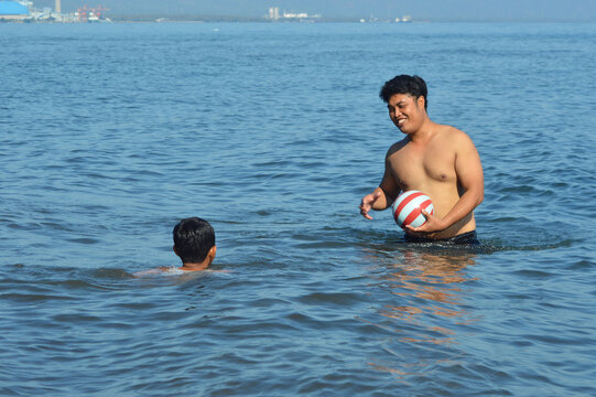 Standing in shallow foamy waves, the father smiles while holding a ball and speaks to his son who swims nearby, creating a warm and playful bonding moment together.