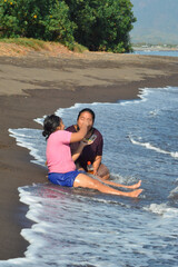 On the wet sand, a smiling daughter feeds her mother a snack while the mother holds a plastic bottle, both enjoying the calm seaside atmosphere with gentle waves.