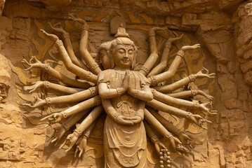 A stone sculpture with many arms stands against a textured wall in China. The figure holds its hands together in a gesture of prayer and contemplation.