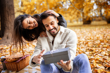 Romantic young couple giving presents to each other in colorful autumn park, joyful and loving fall moment.