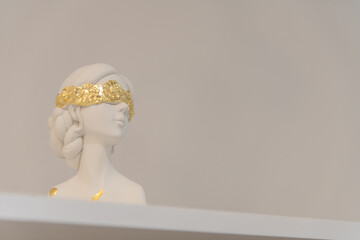 White sculpture of a woman with golden blindfold sits on a shelf