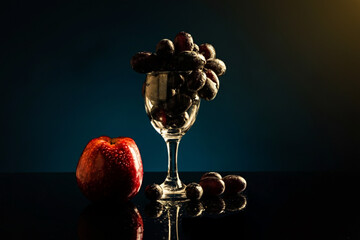 A wine glass filled with black grapes. A red apple kept at the base of wine glass. Few grapes are...