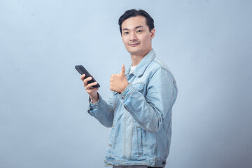 Fototapeta premium Asian man in denim jacket holding smartphone and showing thumbs up gesture with confident smile on plain studio background