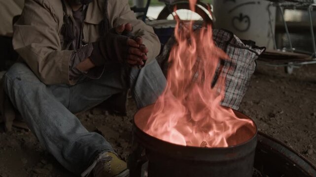 Cropped shot of bearded middle-aged vagrant dressed in jacket and jeans warming and rubbing hands sitting on ground near steel barrel with fire burning