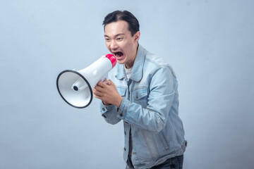 Asian man in denim jacket shouting through a white megaphone with an expressive face, symbolizing...