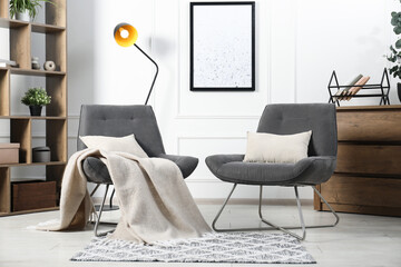Stylish armchairs with pillows, blanket, dresser and shelving unit indoors. Interior design