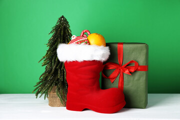 Happy Saint Nicholas day. Red boot with sweets, artificial fir tree and gift box on white wooden table against green background