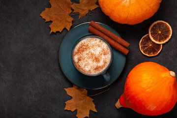 Flat lay of a cozy autumn drink scene with a pumpkin spice latte in a dark ceramic cup, cinnamon sticks, pumpkins, dried oranges, and maple leaves on a dark textured background.