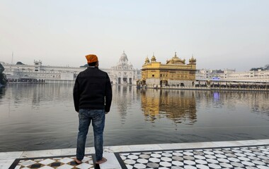 A man wearing a black jacket and orange head wrap handkerchief stands at the edge of the holy pond, gazing at the Golden Temple in Amritsar, Punjab, India.sacred shrine is reflected in the calm water