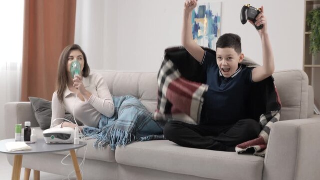 Young woman holding a mask nebulizer and inhaling fumes medication into lungs. Sick lady inhaling through inhaler mask at home and funny boy with gamepad playing video game at home near her