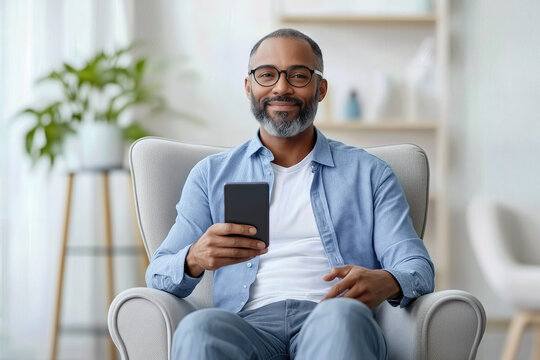 African american man with a gray beard and glasses sitting comfortably in an armchair, smiling while holding a smartphone and enjoying leisure time in a bright living room